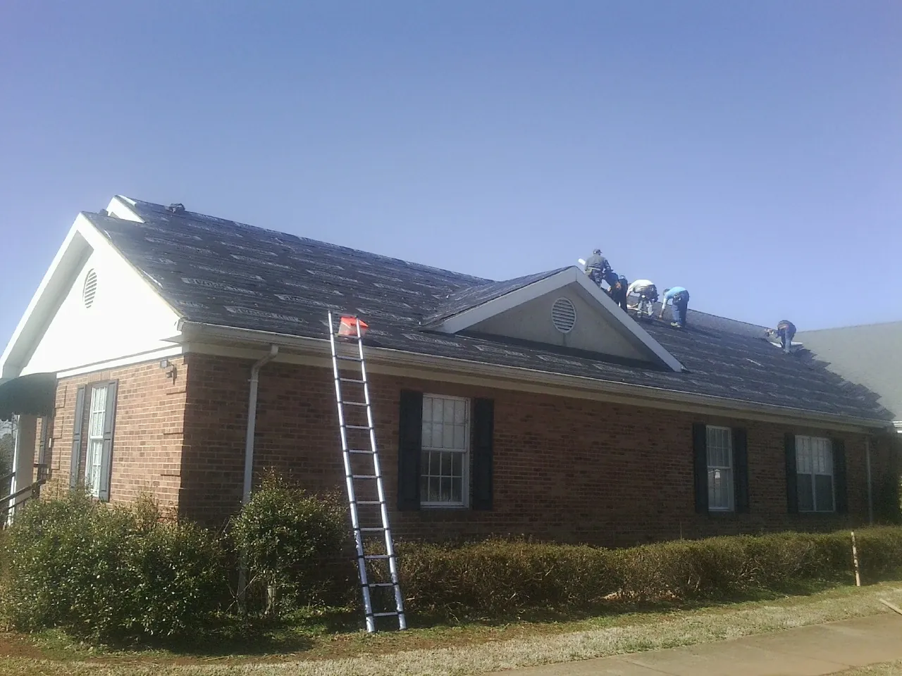 Crew tearing shingles off a brick ranch roof with a ladder set — Metal Roof Repair work by our crew in Junction City